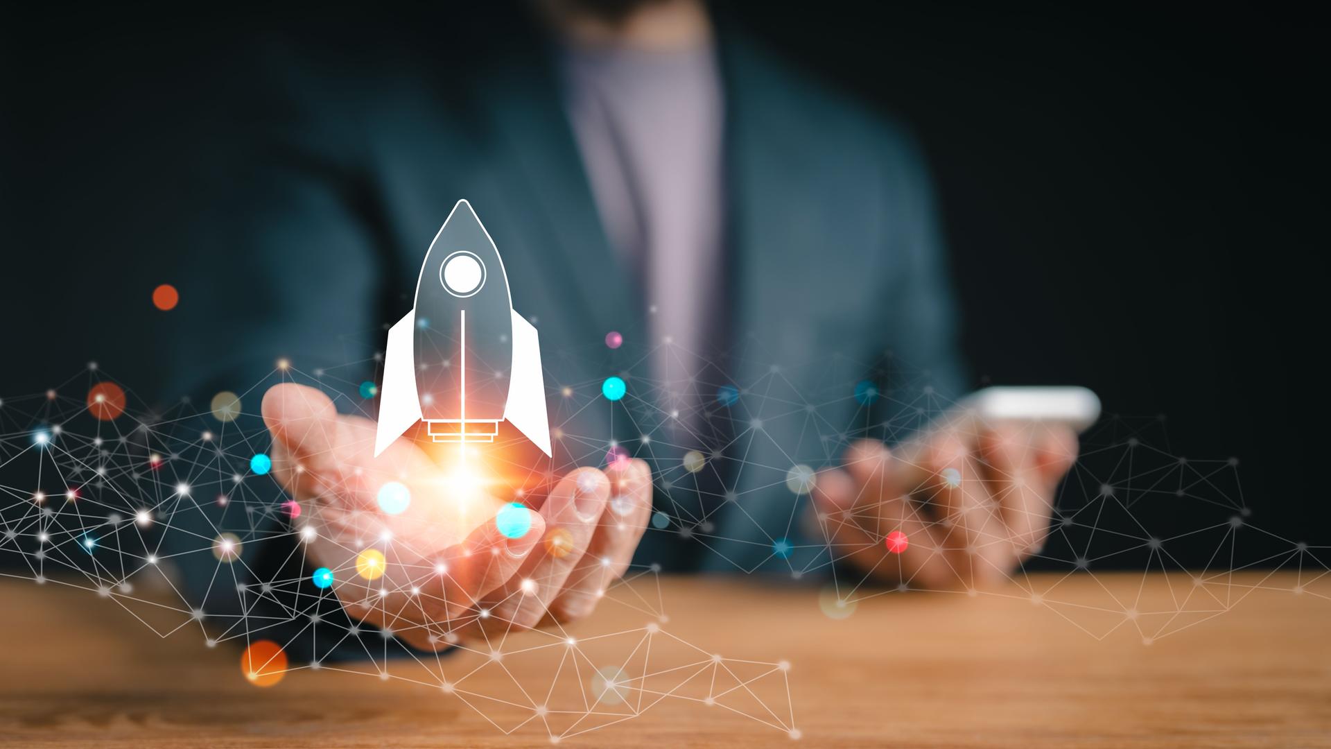 Startup company concept: A businessman using a laptop is shown against a modern, white background with a translucent rocket symbol blasting off and shooting out of the screen.